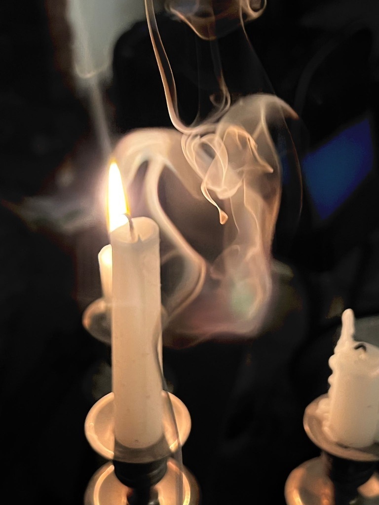 A photograph of candles one lit waiting to be lit and smoke from an extinguished candle